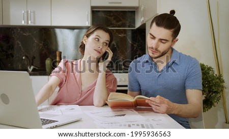 A young woman is talking on the phone while working and her husband is reading a book