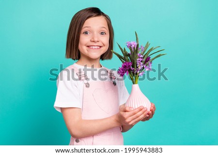 Photo of adorable shiny school girl wear pink overall smiling holding flower vase isolated teal color background