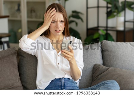 Disappointed young caucasian millennial woman sitting on couch feel frustrated by unexpected bad news on smartphone from bank or cyberbullying concept Royalty-Free Stock Photo #1996038143