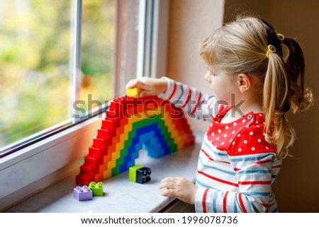 Cute little toddler girl by window create rainbow with colorful plastic blocks. People with rainbows around the world as protest and sign.