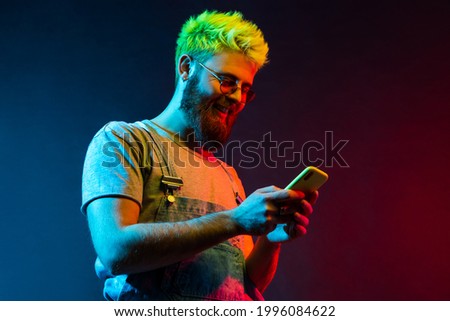 Side view of smiling handsome man with beard and green hair holding smart phone and looking at screen with happy expression, pleasant communication. Colorful neon light, indoor studio shot.