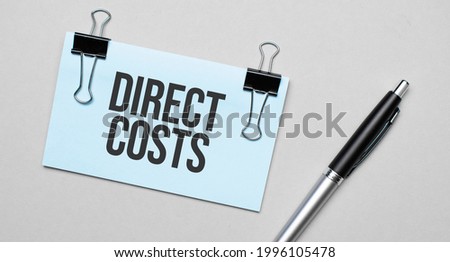Top view of a business card with text DIRECT COSTS , pen, paper clips on a colored background. Business concept.