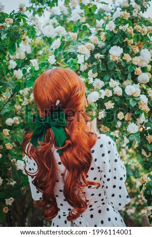 A girl with long red hair stands against the backdrop of a blooming garden. Back View. Women's hairstyle with a green bow. Hair ribbon.