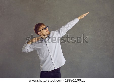 Happy eccentric young guy in 8 bit thug life glasses dancing dub and having fun. Studio portrait business man in 8bit pixel sunglasses doing dab arm move standing against concrete grey wall background