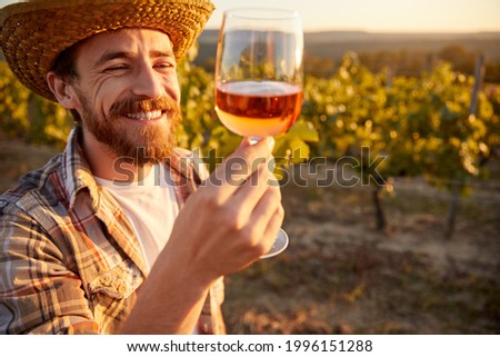 Cheerful bearded male farmer tasting rose wine from glass while standing near grape trees in vineyard Royalty-Free Stock Photo #1996151288