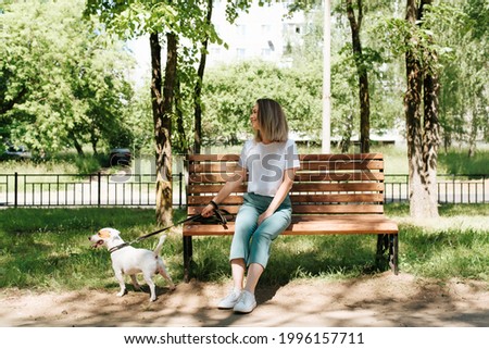 Pretty young woman in stylish clothes resting on a bench while walking the dog. Happy blonde girl and jack russell terrier on a walk in the park on a summer sunny day. Royalty-Free Stock Photo #1996157711