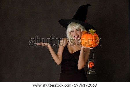 Young Asian female in blond wig and witch hat with Halloween pumpkin lantern laughing happily during party on black background