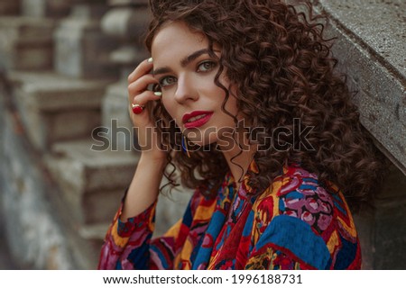 Fashionable woman with long curly hair, red lips makeup, wearing trendy summer colorful printed shirt, stylish ring with big gem, posing in city. Copy, empty space for text Royalty-Free Stock Photo #1996188731