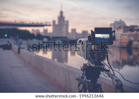 A professional video camera stands on a tripod recording the city and the river at dawn
