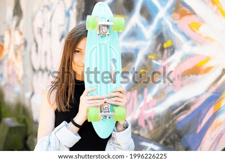 A young beautiful woman in a black sports top and a blue denim shirt covers half of her face with her turquoise skate while standing against a graffiti wall background.