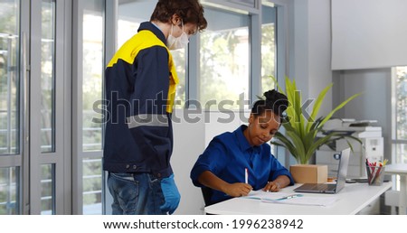 African female consumer signing document to receive parcel from courier in protective mask. Postman in safety mask and gloves deliver package for afro-american businesswoman in office