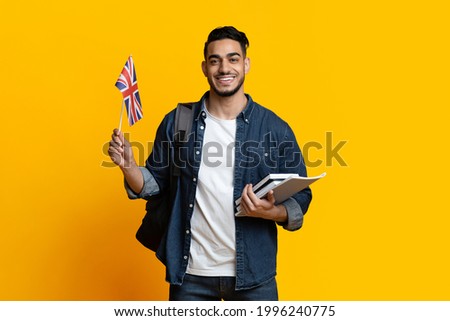 Smiling arab guy student with backpack and bunch of books showing flag of Great Britain over yellow studio background, copy space. Positive middle-eastern young man studying English at school Royalty-Free Stock Photo #1996240775