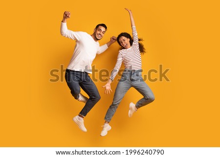Cheerful Active Arab Couple Jumping In Air Over Yellow Background, Joyful Emotional Middle Eastern Man And Woman Having Fun Together, Raising Hands And Exclaiming With Excitement, Full Length Royalty-Free Stock Photo #1996240790