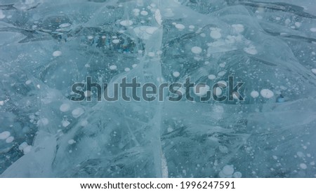 Gently ice blue. Close-up. Full frame. In detail. On the shiny surface, there is a natural pattern of intersecting deep cracks and bubbles of frozen methane gas. Baikal