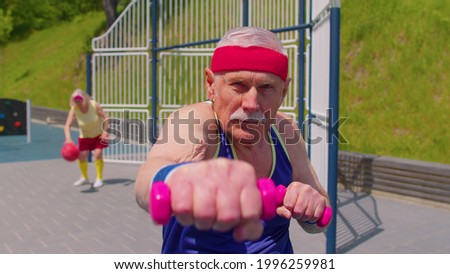 Active senior handsome man grandfather doing sport training boxing fitness aerobics cardio exercising with dumbbells on basketball playground. Healthy grandmother playing ball basketball on background