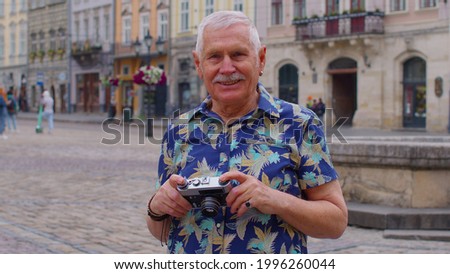 Portrait of senior grandfather man tourist taking pictures with photo camera, smiling using retro device in summer city center. Photography, travelling, vacation. Active grandpa life after retirement