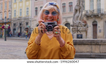Portrait of senior woman tourist taking pictures with photo camera, smiling using retro device in summer city center of Lviv, Ukraine. Active life after retirement. Photography, travelling, vacation.
