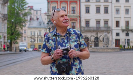Senior man tourist with retro photo camera, smiling, listening music on earphones, dancing on summer city street center of Lviv, Ukraine. Photography, travelling vacation. Active life after retirement