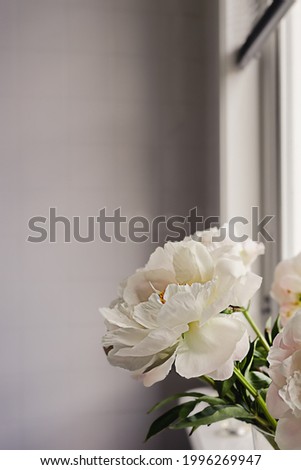 White peony flowers bouquet with green stems and leaves on a blurred tile wall background