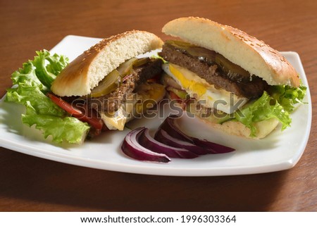 Large delicious hamburger cut with fried patty, fried egg, pickles, tomato and lettuce on a white plate