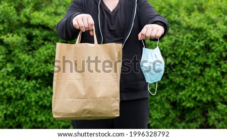 Close up of man hand with mask and paper bag for takeaway food on nature green background. Delivery in any weather around the clock to the client.