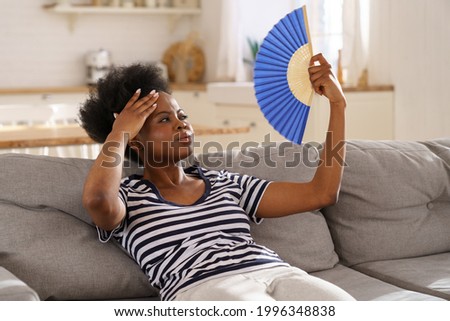 Tired African American woman suffering from heatstroke flat without air-conditioner, waving blue fan, sitting on sofa at home. Black girl cooling in hot summer weather. Overheating, high temperature. Royalty-Free Stock Photo #1996348838