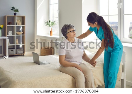 Trying to make patient's life happier. Female home care nurse supports and assists senior woman with all her daily needs. Caregiver holds old lady's hand, helps her stand up from bed and talks to her Royalty-Free Stock Photo #1996370804