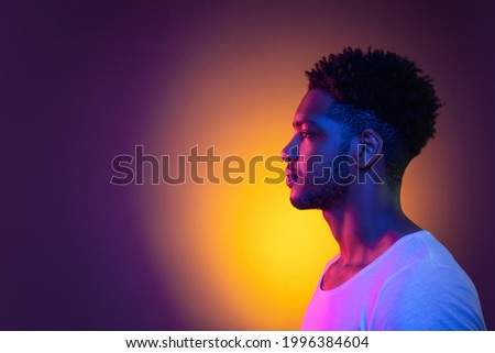 Portrait of a young pensive african man in profile at studio. Male model isolated in colorful bright neon lights posing on purple background. Side view.