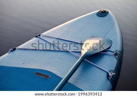 Paddle board and surf board with paddle on blue water surface background close up. Surfing and SUP boarding equipment in sunset lights close-up. Outdoor water sports. Surfing lifestyle backgrounds. Royalty-Free Stock Photo #1996393412