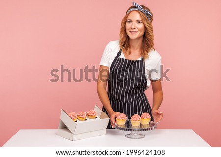 Young adult female baker serving cakes, wearing white T-shirt and black striped apron, looking at camera with satisfied facial expression. Indoor studio shot isolated on pink background.