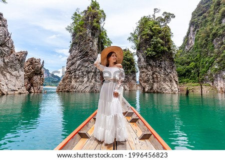 Happy Young Female Tourist in Dress and Hat at Longtail Boat near Famous Three rocks with Limestone Cliffs at Cheow Lan Lake. Travel Woman Standing on Boat in Khao Sok National Park in Thailand Royalty-Free Stock Photo #1996456823