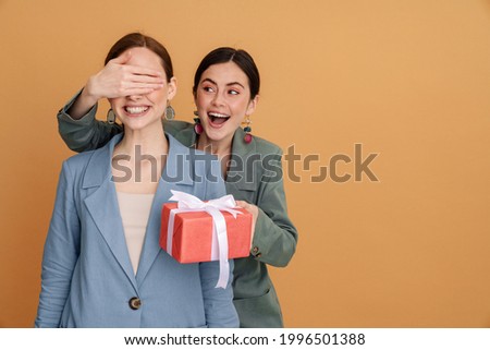 Young woman covering her friend's eyes while giving present isolated over yellow background