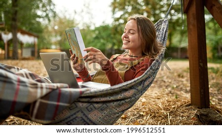 young woman is reading her notes while lies in a hammock, she is holding a laptop on her legs and smiling, woman working remotely on the fresh air 