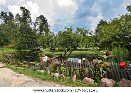 View on a little lake, surrounded by the trees, forest in the background; old, damaged fence with some pots on it and rusted bike on the foreground