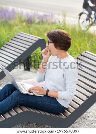 Woman sits with laptop on urban park bench. Freelancer yawns at work. Student learns remotely from outdoors. Modern lifestyle.