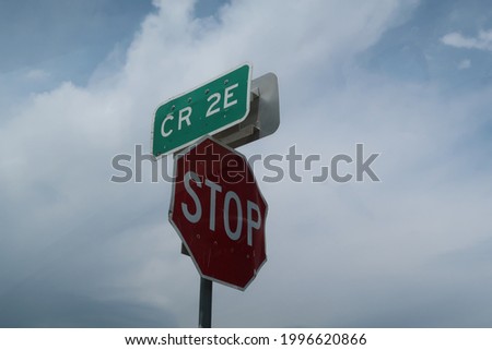 Green street sign for county road 2E and a stop sign against a cloudy blue sky