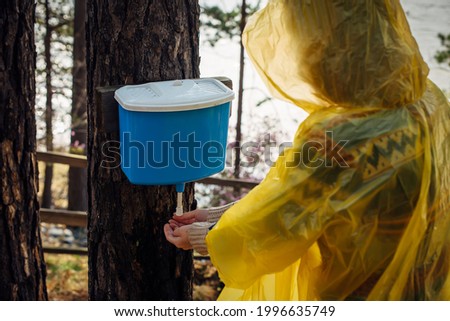 Unknown woman in yellow raincoat washes her hands in wash basin hanging on tree. Rainy morning on tourist camp in forest near the river.