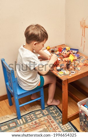 Blond little boy in white t-shirt draws picture with brush and brown color at table with dirty jars and toys in children room