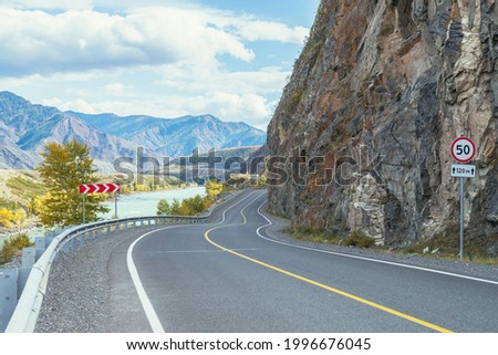 Vivid autumn landscape with mountain highway along big mountain river in sunshine. Bright alpine scenery with wide turquoise river and mountain road in autumn colors. Highway in mountains in fall time