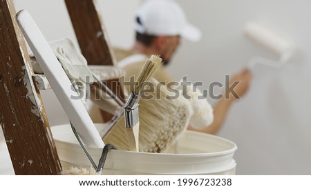 hands of house painter man decorator work of home to renovate, using roller paint, white bucket on wooden ladder with paint brushes as background, close-up Royalty-Free Stock Photo #1996723238