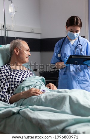 Medical assistant with protection mask checking treatment of senior man reading notes on tablet pc. Sick elderly laying in bed discussing with nurse about diagnosis expertise with iv bag attached.