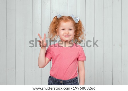 Portrait of cute carefree ginger little girl 4-6 years old, showing peace v-sign gesture. Studio shot, light wooden background