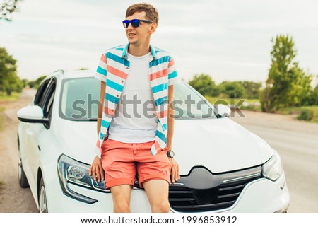 Happy young man, in sunglasses, in summer clothes, standing near his car, enjoying the sunset of summer nature, travel concept