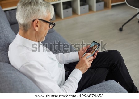 Concept of older adults using online dating apps or websites. Old bachelor searching for future wife. Senior man holding mobile phone in hand and looking at photo of beautiful young woman on display