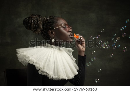 Blowing bubbles. Medieval African young woman in black vintage dress with big white collar isolated on dark green background. Concept of comparison of eras