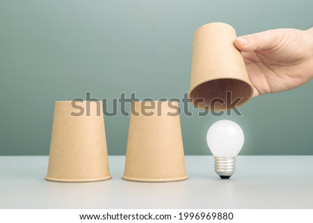 Hand reveal the right cup in shell game and found the glowing light bulb, finding good idea and success solution, chance and making a decision concept Royalty-Free Stock Photo #1996969880