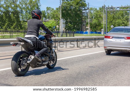 A female biker in dark clothes is moving on a modern sports motorcycle behind a passenger car from left to right on the highway in urban conditions during the day.