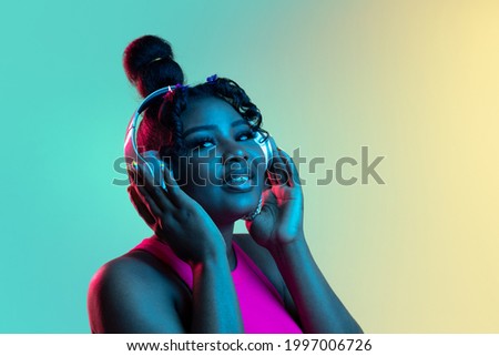 Joyful young beautiful African girl listensng to music in headphones isolated on blue green background in neon light. Concept of human emotion, facial expression. Copy space for ad.