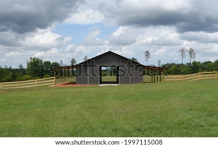 Picture perfect image of post frame horse barn in green pasture under cloudy sky with surrounding wooden fence 