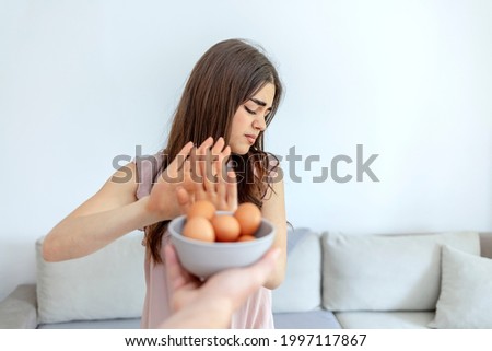 Photo of a woman refusing to eat eggs. Egg free affected allergy banned restriction. Young beautiful girl holding fresh egg at home with open hand doing stop sign with serious and confident expression